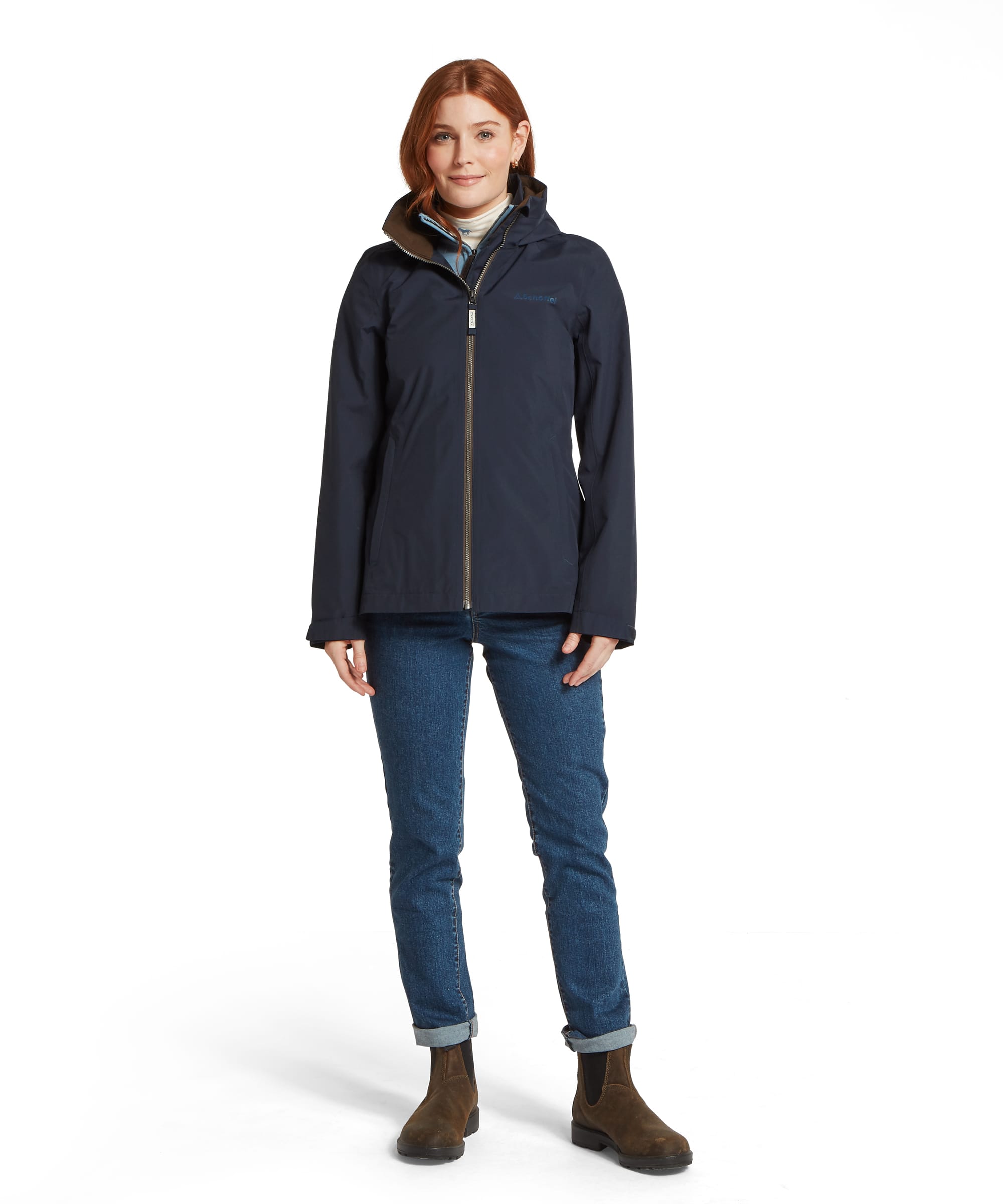 Schöffel Women's Manton Lightweight Waterproof Jacket in navy, full-length front view with storm hood, gold-tone zip, side hand-warmer pockets and relaxed fit, layered over a blue gilet and cream roll-neck, worn with blue jeans and tan suede Chelsea boots