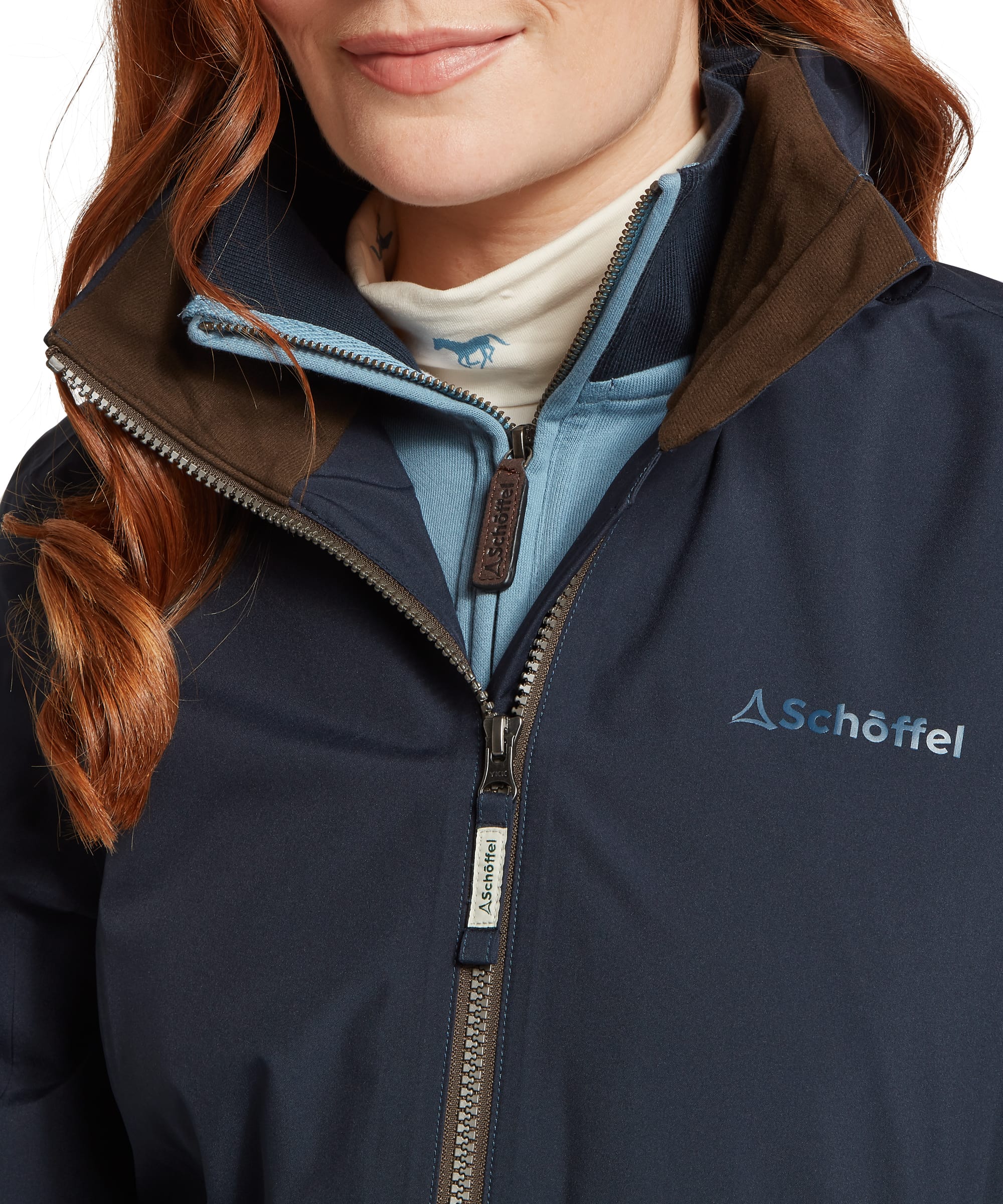 Close-up of the storm collar, zip fastening and Schöffel branding on the Women's Manton Lightweight Waterproof Jacket in Navy, showing the contrast brown suede-look trim and layering over a blue quarter-zip fleece and cream neck gaiter