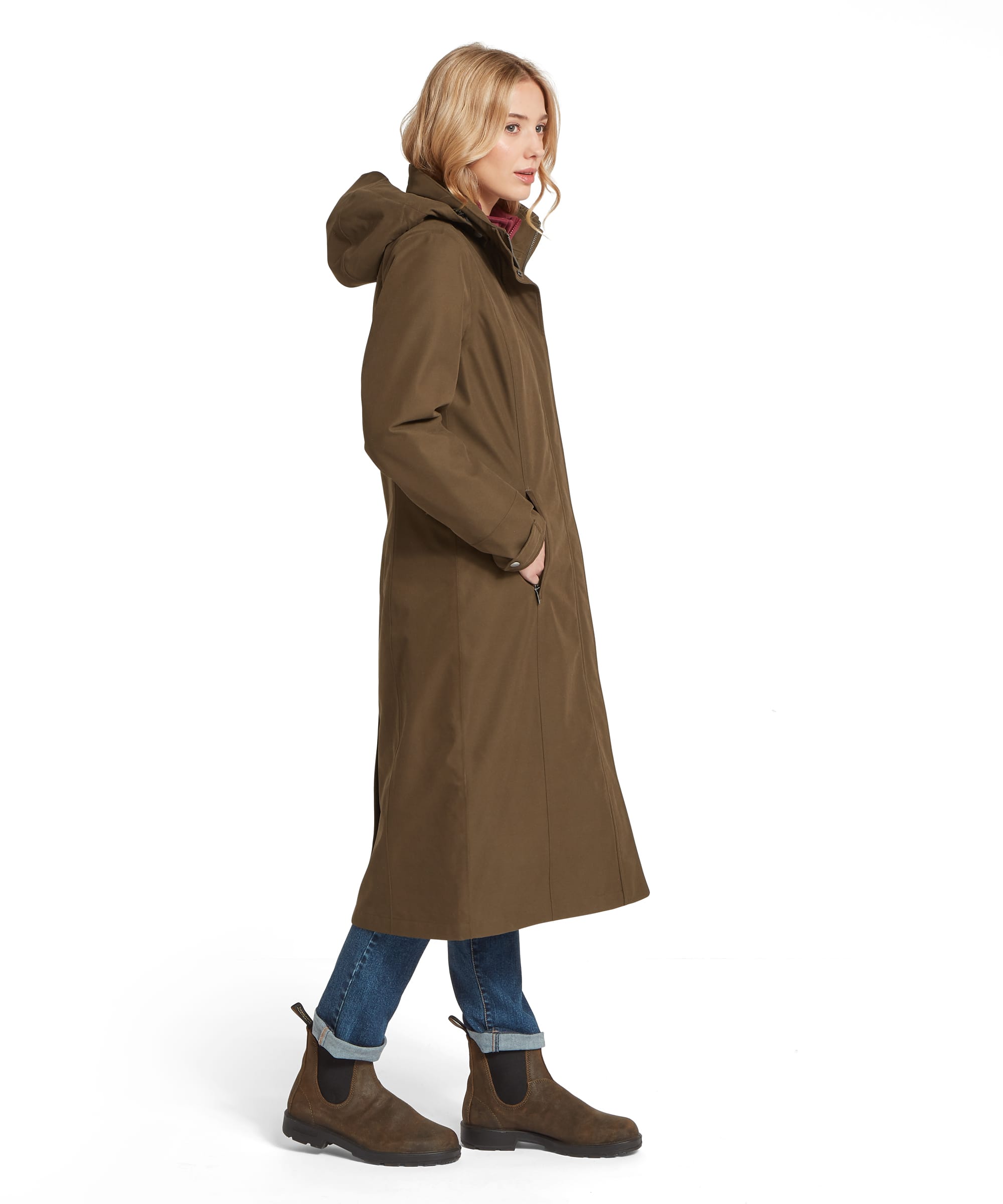 Side profile of a woman wearing the Schöffel Women's Marefield Waterproof Coat in Dark Olive, showing the full-length silhouette, structured hood, zip fastening and A-line shape, styled with turn-up blue jeans and suede Chelsea boots
