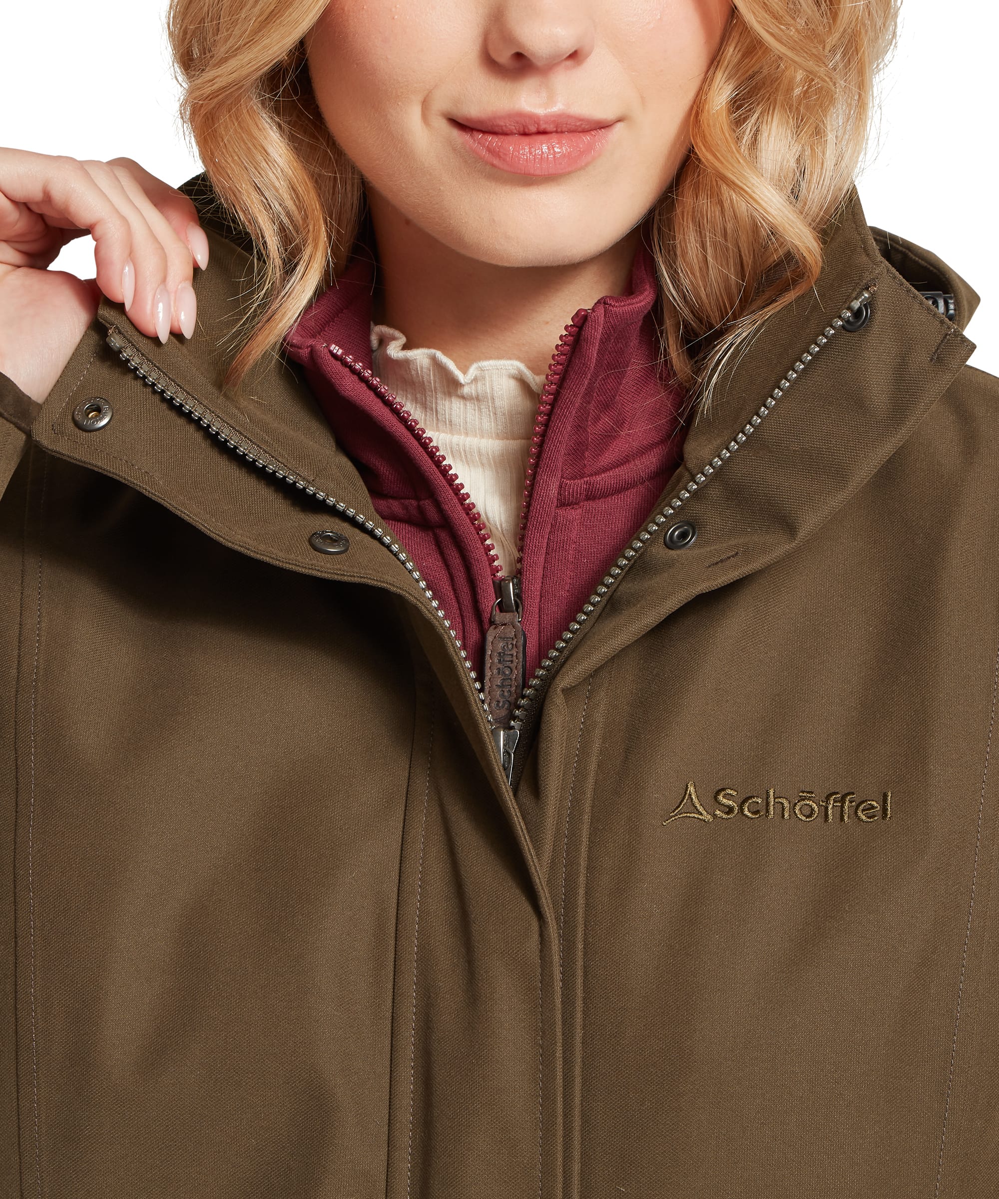 Close-up of the storm collar, Schöffel branded zip pull and tonal embroidered logo on the Women's Marefield Waterproof Coat in Dark Olive, with layering over a burgundy quarter-zip fleece and cream polo neck visible underneath