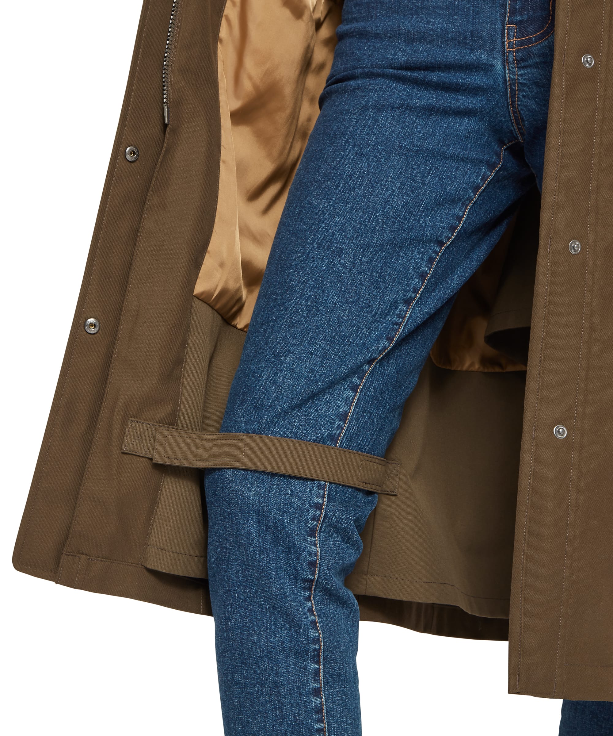 Interior hem detail of the Schöffel Women's Marefield Waterproof Coat in khaki green, showing the open coat revealing the warm caramel satin lining, press-stud vent adjusters at the hem, and an interior tab loop, styled with blue denim jeans