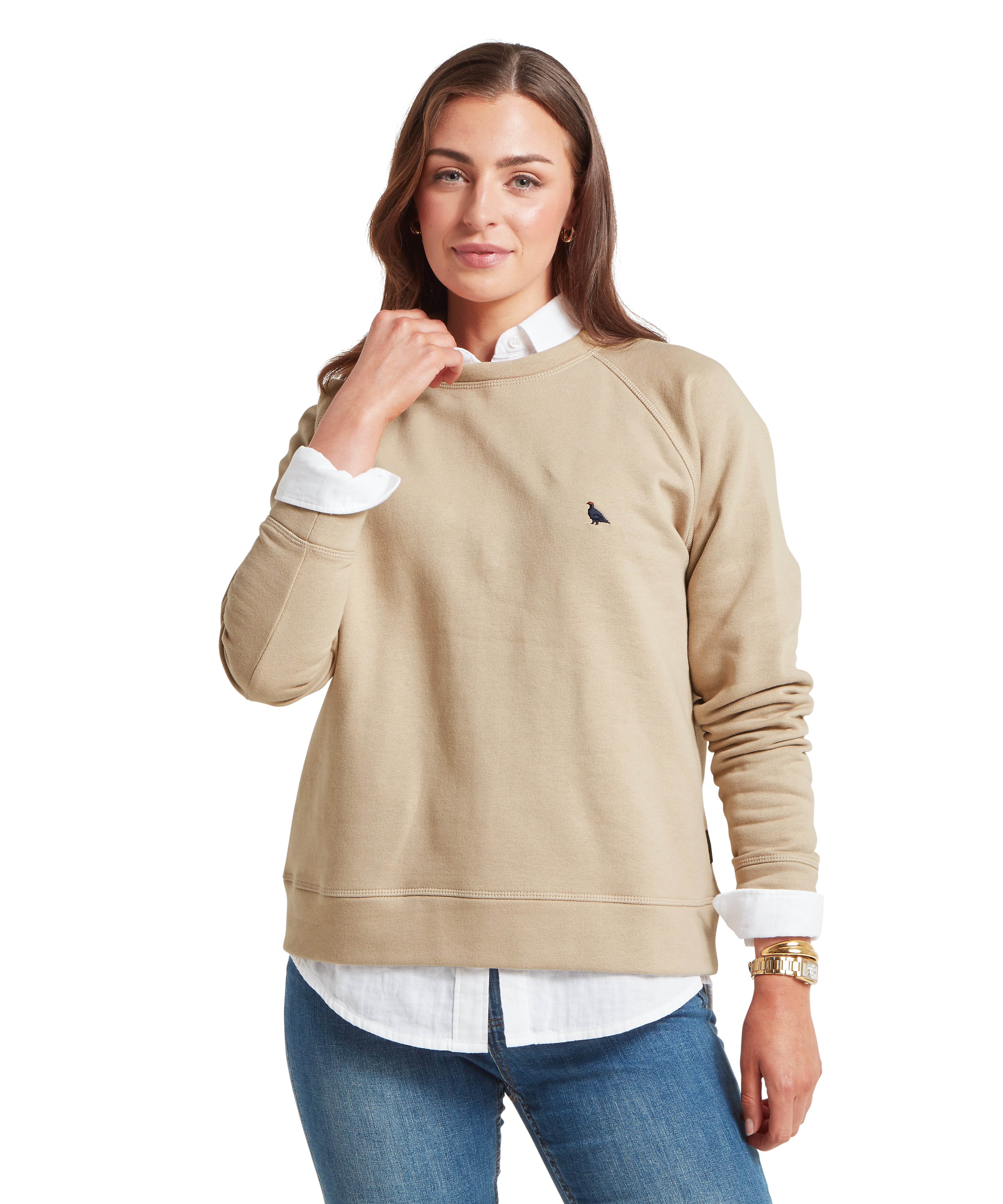 Woman wearing a beige sweatshirt with a small ptarmigan logo, white shirt, and blue jeans on a white background