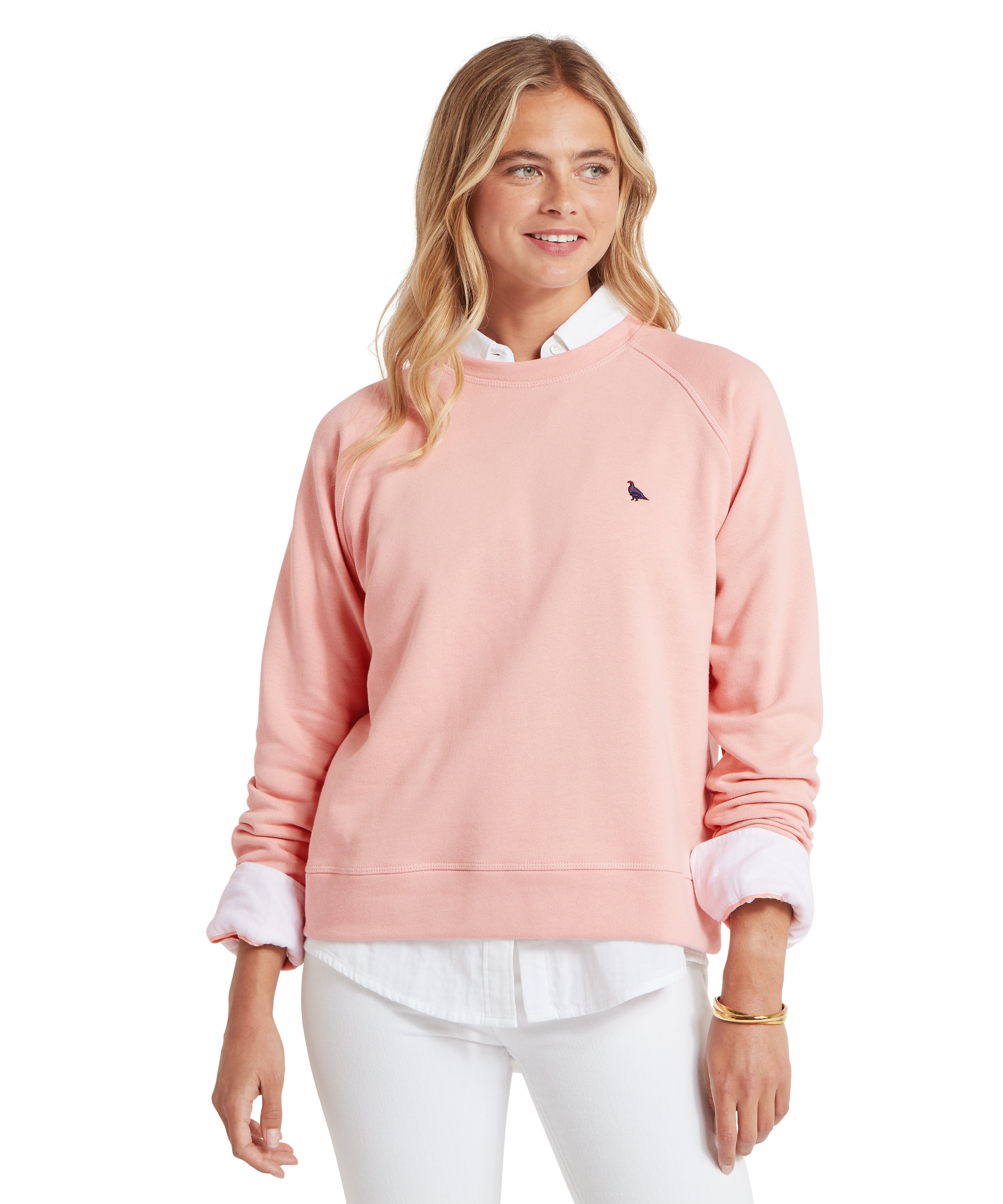 Woman wearing a pink sweatshirt with a small  ptarmigan bird logo, white shirt, and white trousers on a white background