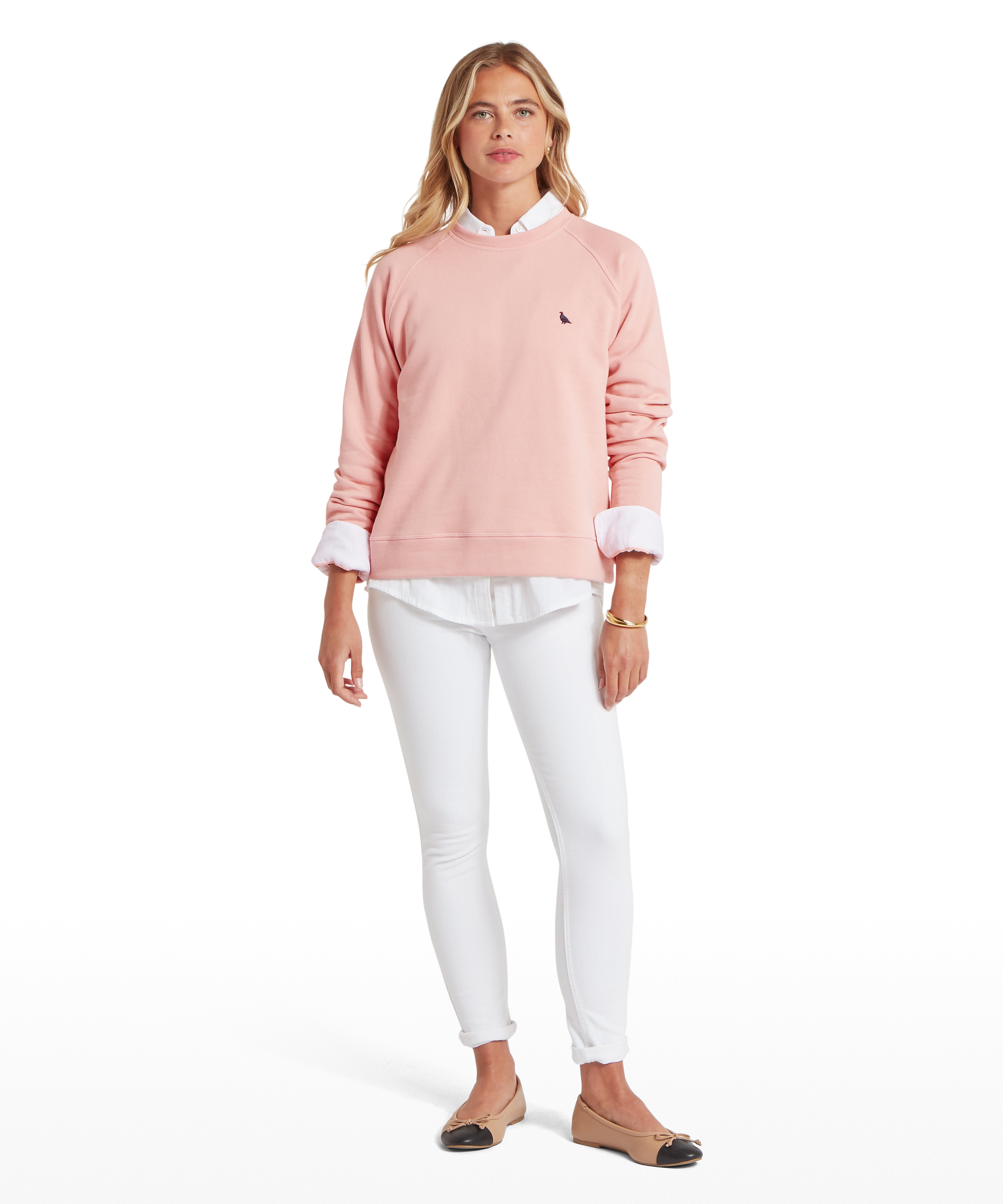 Person standing and facing forward wearing a light pink sweatshirt with a small embroidered ptarmigan bird logo on the chest, layered over a white button-down shirt with visible collar and cuffs. Paired with fitted white jeans and beige flats with black toe caps. The individual has long, loose hair and wears a gold bracelet on the left wrist, posed against a plain white background.