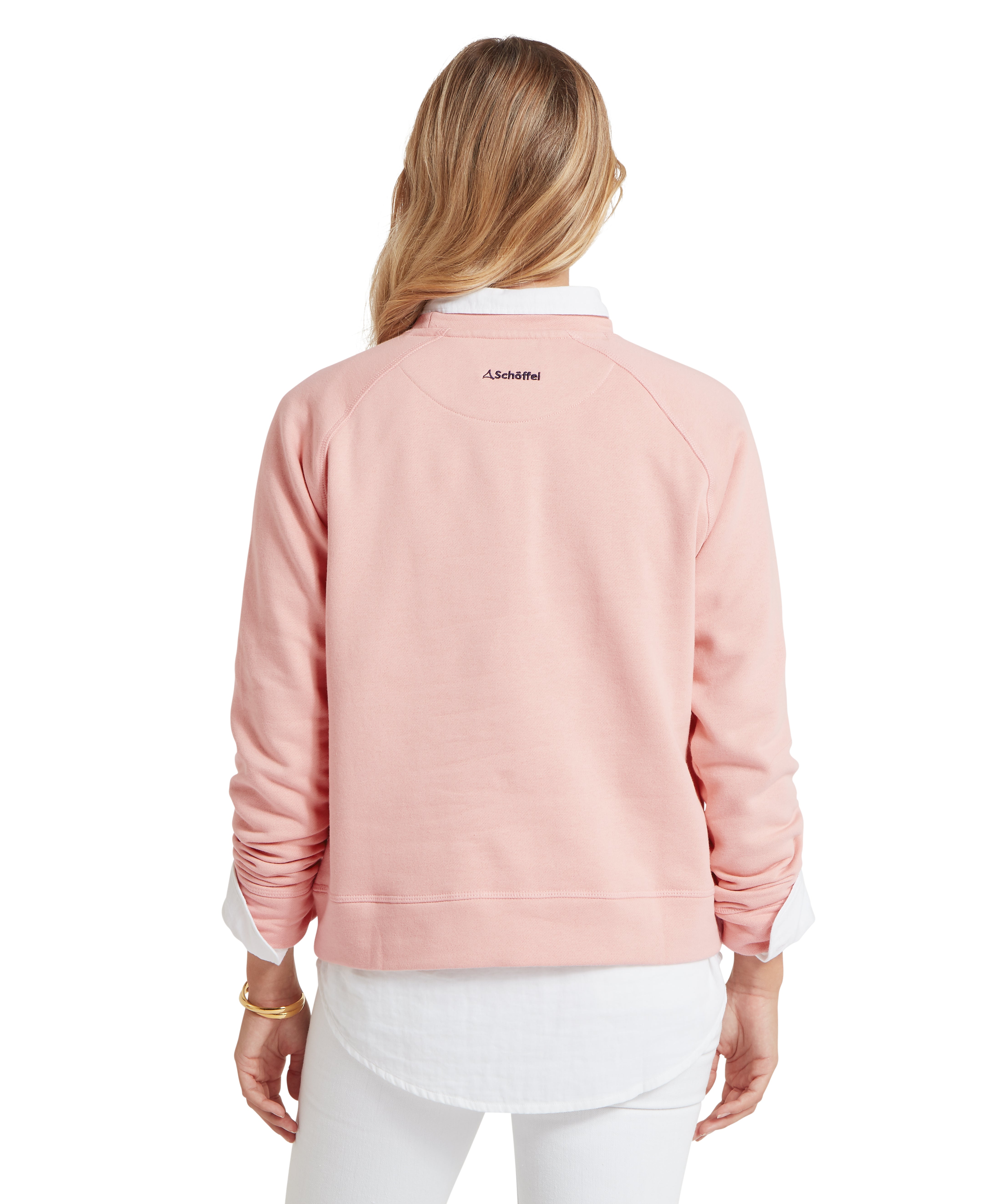 Back view of a person wearing a light pink long-sleeve sweatshirt layered over a white collared shirt, with rolled-up sleeves and small embroidered text "Schöffel" near the neckline. The individual has long, wavy hair and is standing against a plain white background.