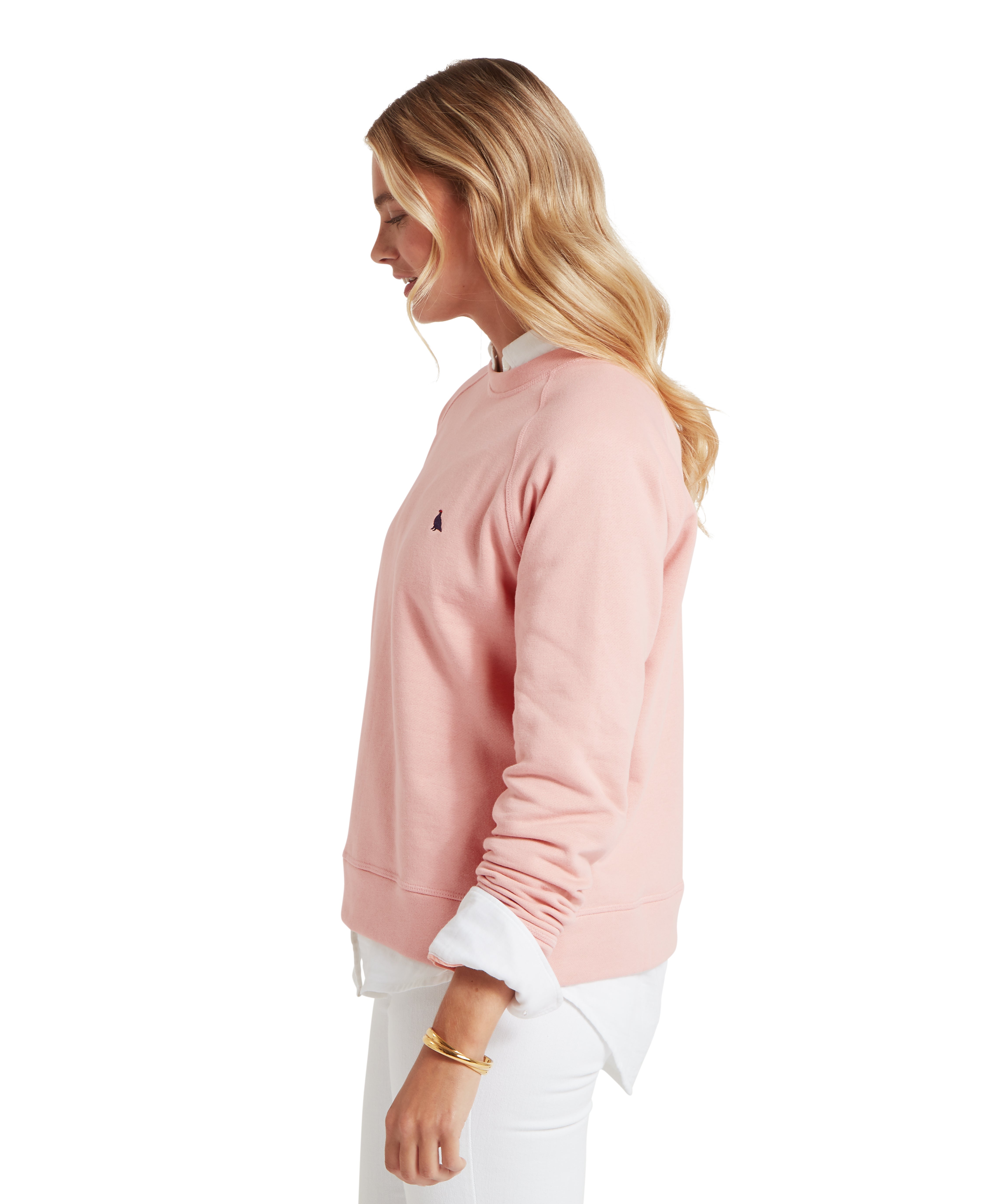 Profile view of a person facing left, wearing a light pink sweatshirt over a white collared shirt with visible cuffs and hem. The sweatshirt features a small dark-coloured ptarmigan logo on the chest. Paired with white trousers and accessorized with a gold bracelet on the left wrist. The individual has long, wavy hair cascading over the shoulder, posed against a plain white background.