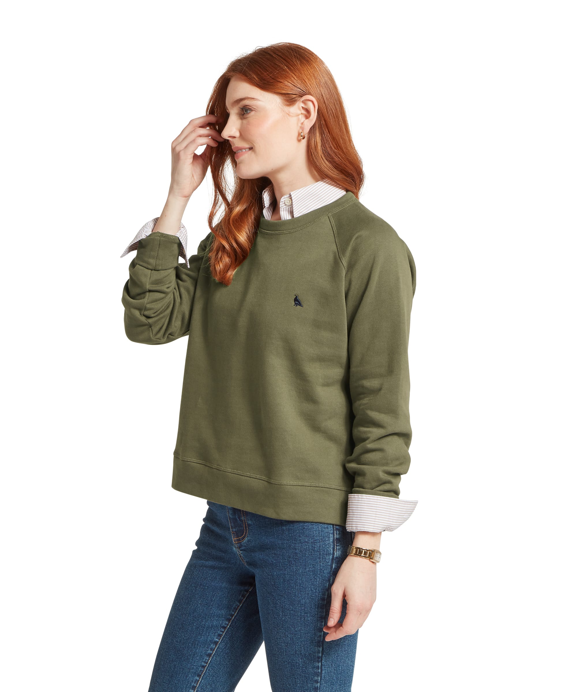 Three-quarter view of a woman wearing the Schöffel Women's Newberry Sweatshirt in Khaki Green, showing the embroidered Schöffel bird logo, raglan seam detail and layering over a classic striped shirt with blue jeans