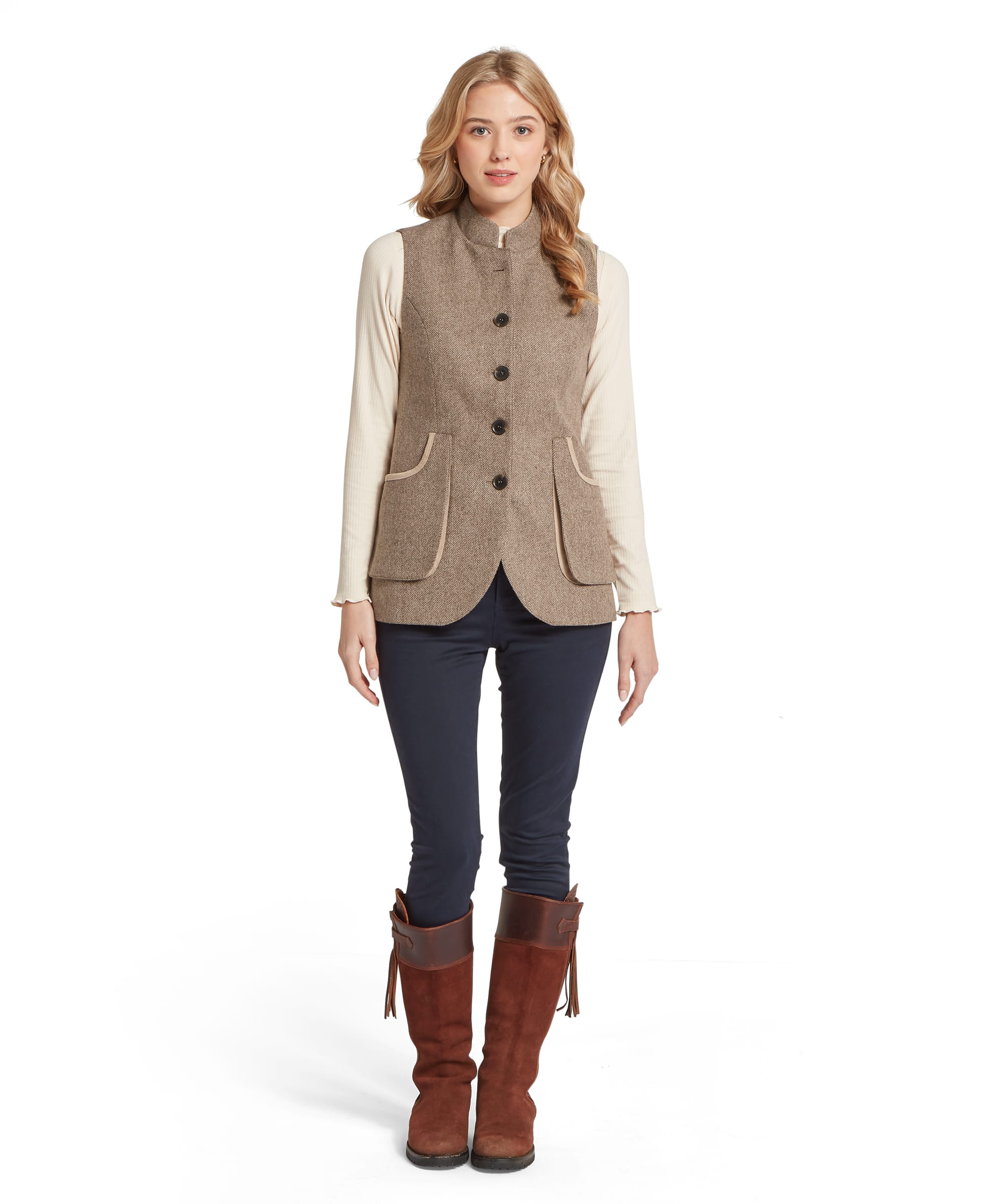Full-length view of the Schöffel Women's Pennan Tweed Nehru Waistcoat in Natural Herringbone, worn over a cream ribbed top and navy trousers with tan suede tassel riding boots, showing the fitted waistcoat silhouette and scalloped hem