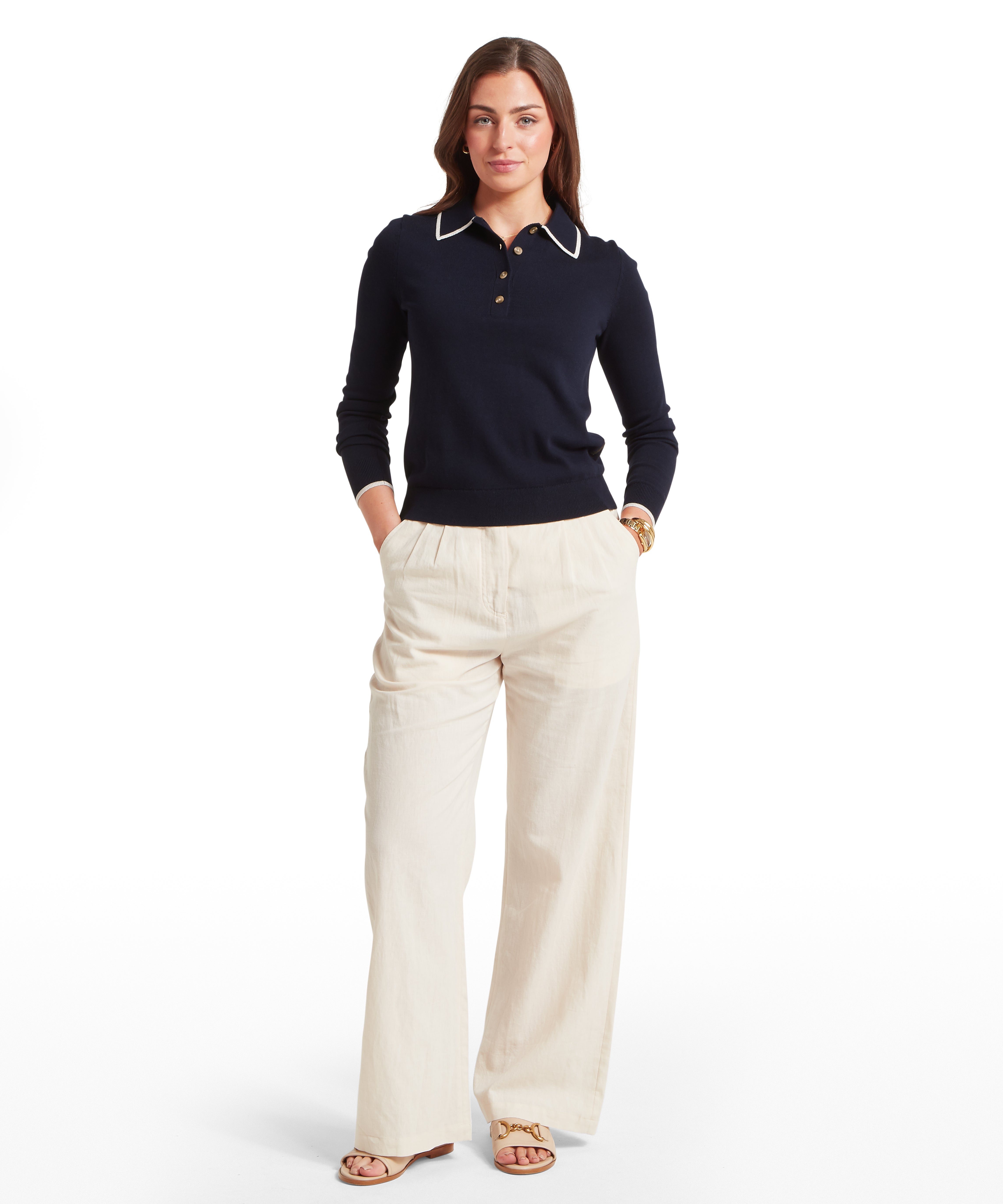 Woman wearing a dark navy long-sleeve knit polo sweatshirt with white contrast trim on the collar and cuffs and a three-button placket, paired with light beige trousers, standing against a white background. The woman is facing forwards and has both hands in her pockets.