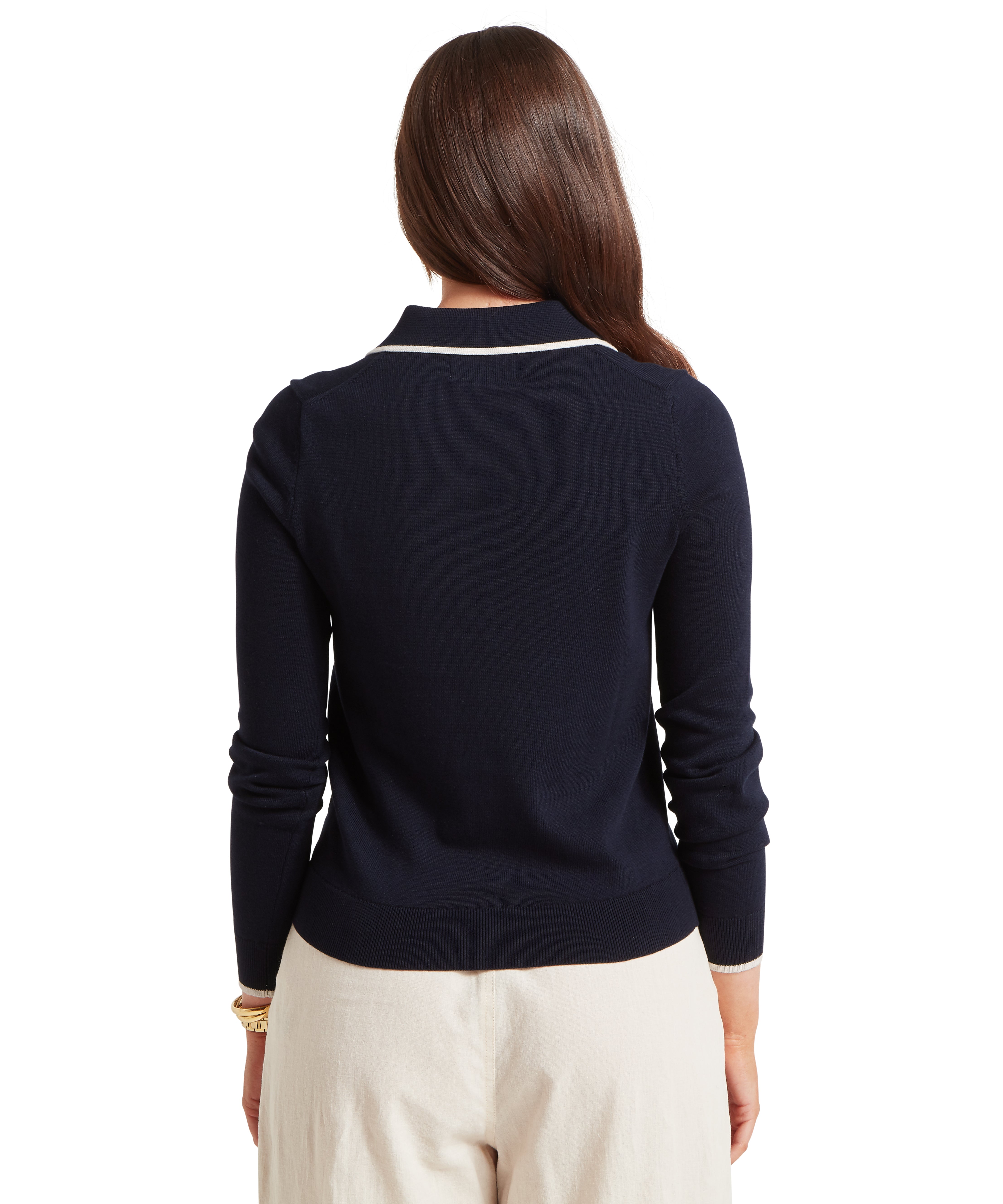 Back view of a person wearing a navy blue jumper with white trim along the collar, paired with cream trousers and a gold bracelet on the left wrist; long brown hair draped over the right shoulder.