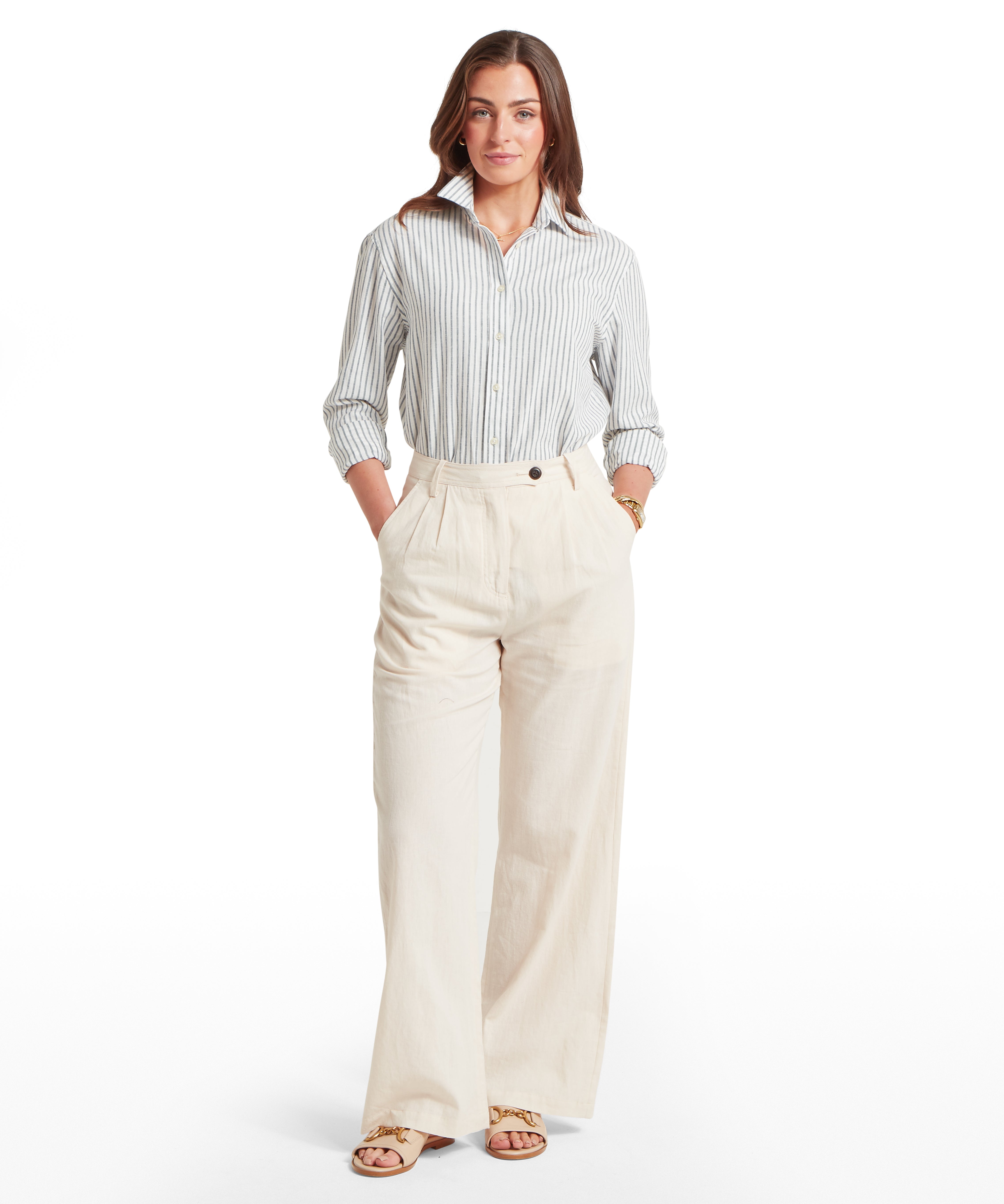 Woman standing with a confident posture, facing forward against a white background while wearing a white and navy blue vertical striped button-down shirt tucked into high-waisted, wide-leg cream trousers. The shirt has its sleeves rolled to the mid-forearm and the collar slightly popped, while the trousers feature pleated front details and are paired with tan open-toe sandals decorated with gold chain hardware.