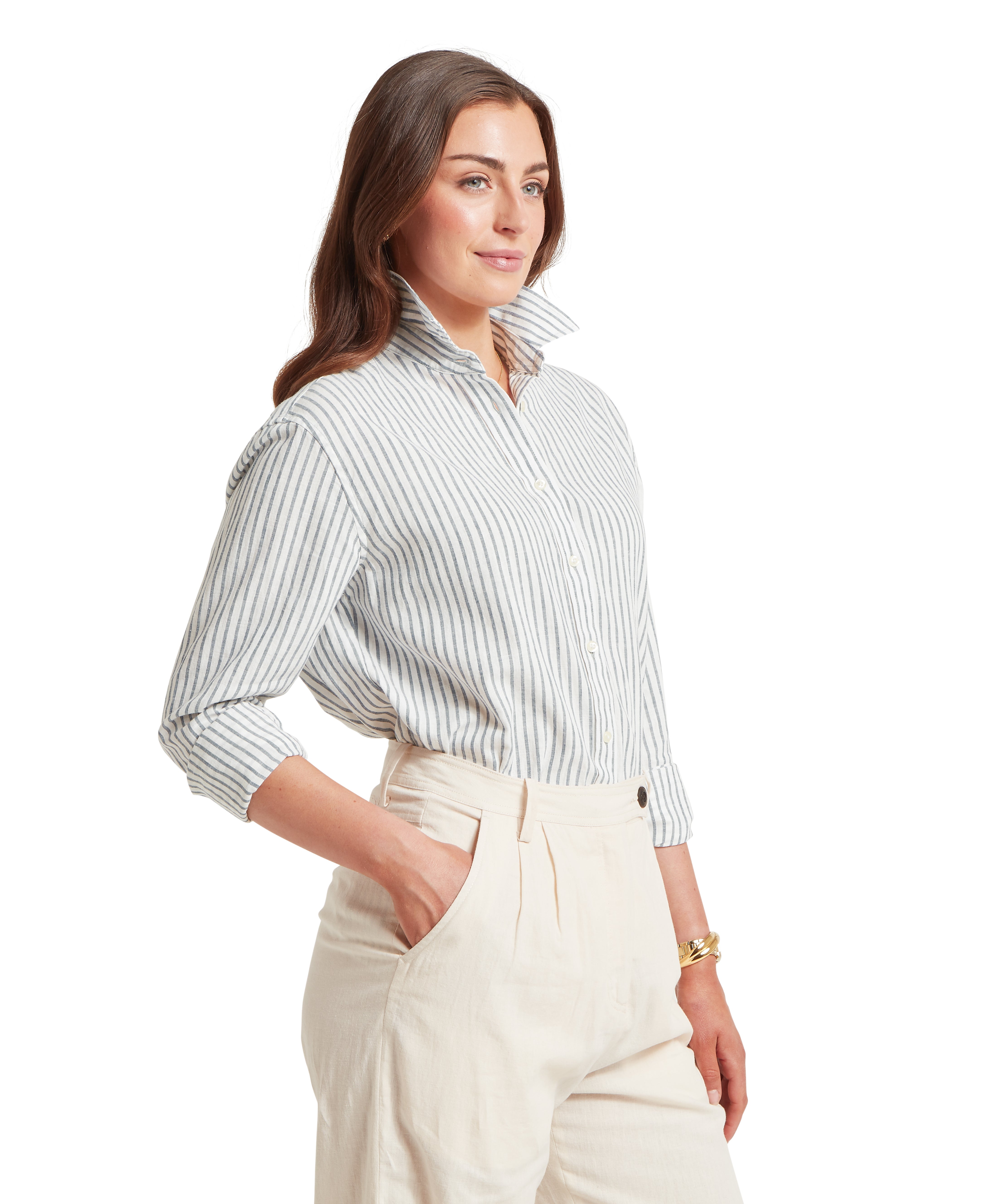 Woman standing in a three-quarter side profile wearing a white and navy blue vertical striped button-down shirt tucked into high-waisted cream trousers. She has her right hand tucked into her pocket, showing the pleated detail of the trousers, and is wearing a gold watch and bracelet on her left wrist. The shirt features a structured, popped collar and sleeves neatly rolled up to