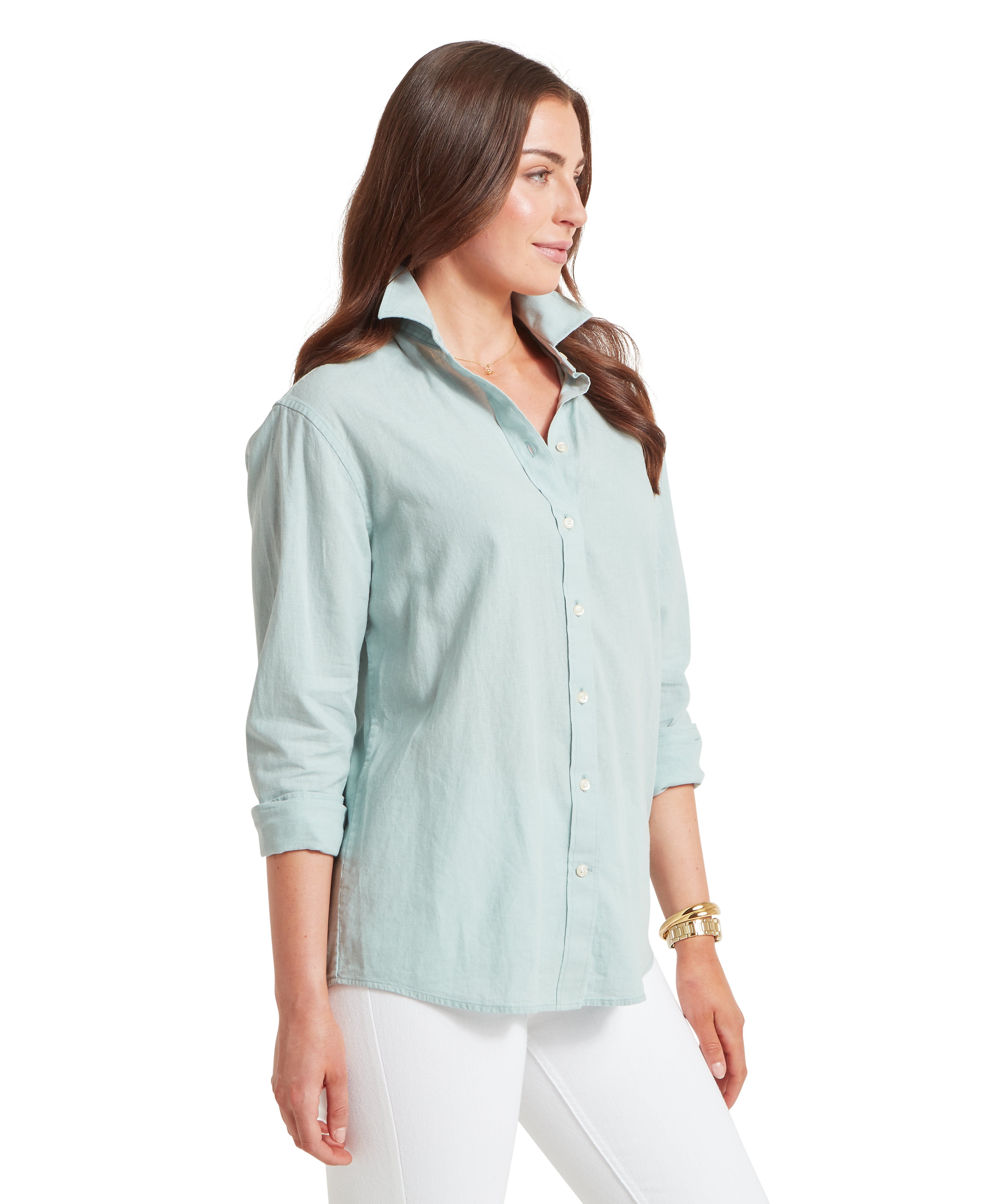 Woman standing in a three-quarter side profile wearing a light mint green button-down shirt with the sleeves rolled up to her forearms. The shirt features a structured, popped collar and small white buttons along the front placket. She is wearing white trousers, a thin gold necklace, and a gold watch and bracelet on her left wrist while her long brown hair falls over her shoulders.