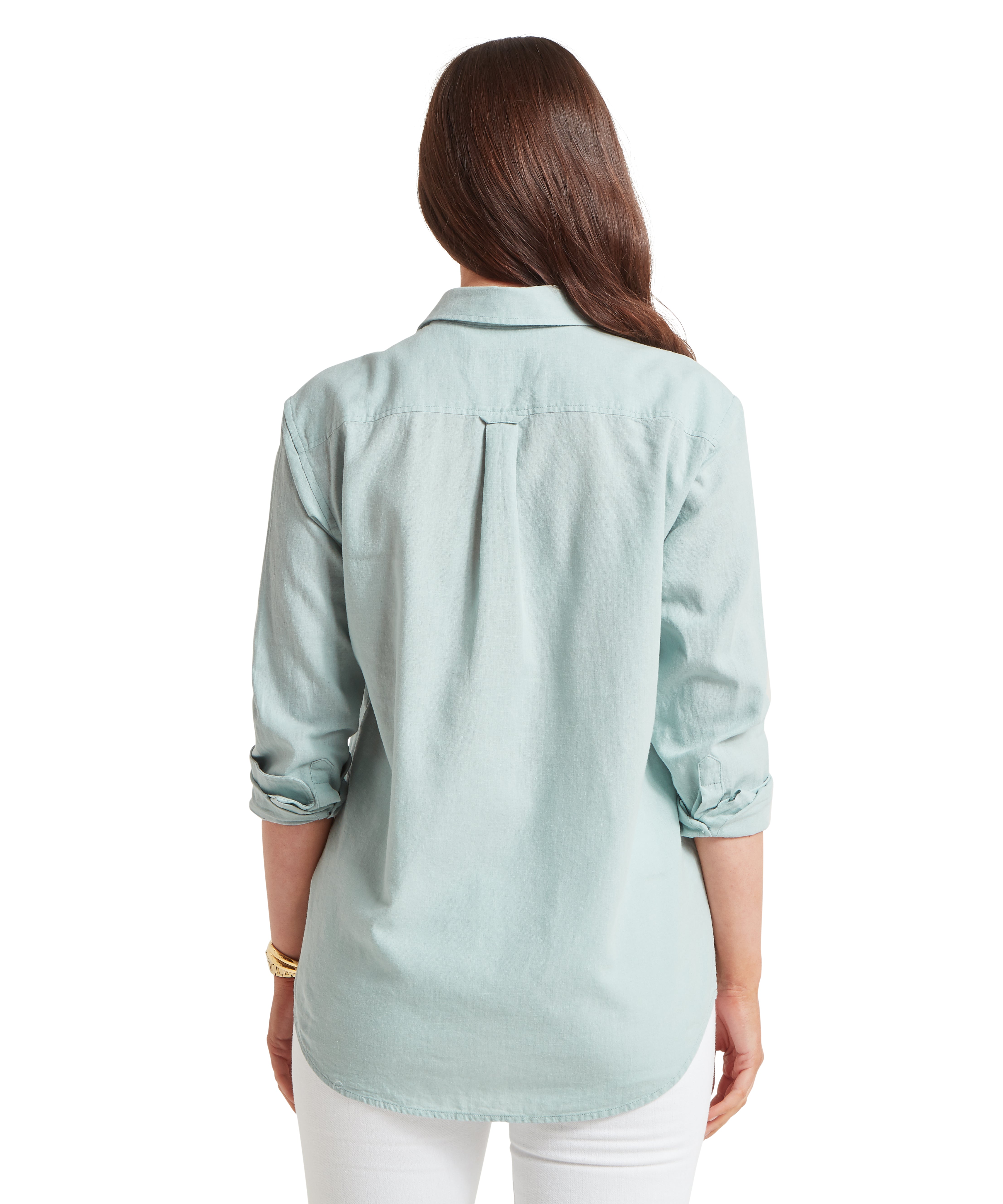 Woman seen from behind with long, dark brown hair, wearing a light mint green linen button-down shirt that features a horizontal yoke across the shoulders and a central box pleat for a relaxed fit. The shirt has a curved hemline that falls over white trousers, and the sleeves are neatly rolled up to the elbows, revealing a gold watch on her left wrist.