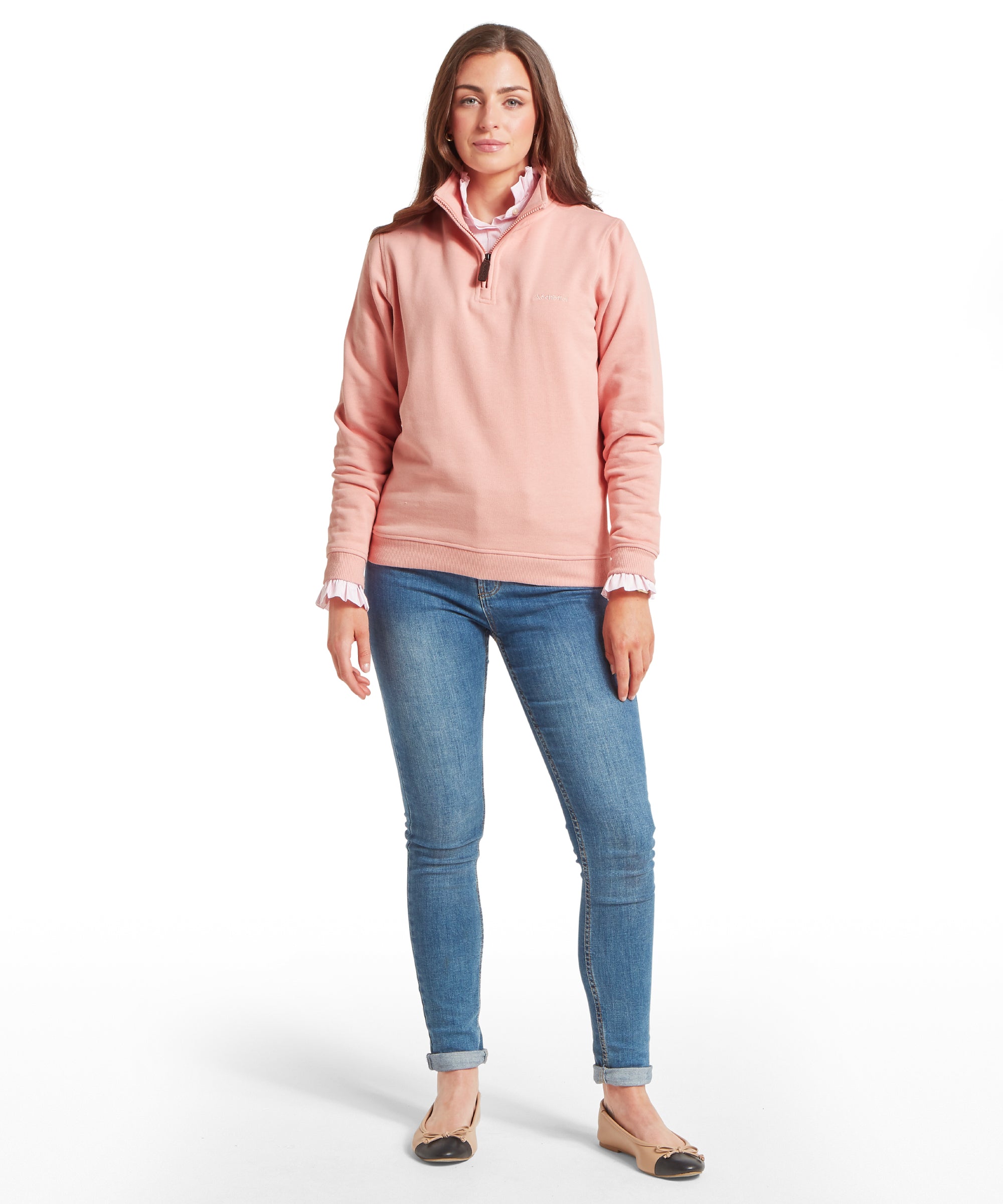 Woman wearing a light pink quarter-zip sweatshirt over a collared shirt, paired with blue skinny jeans. Styled with beige flats featuring black toe caps. Standing front-facing against a plain white background