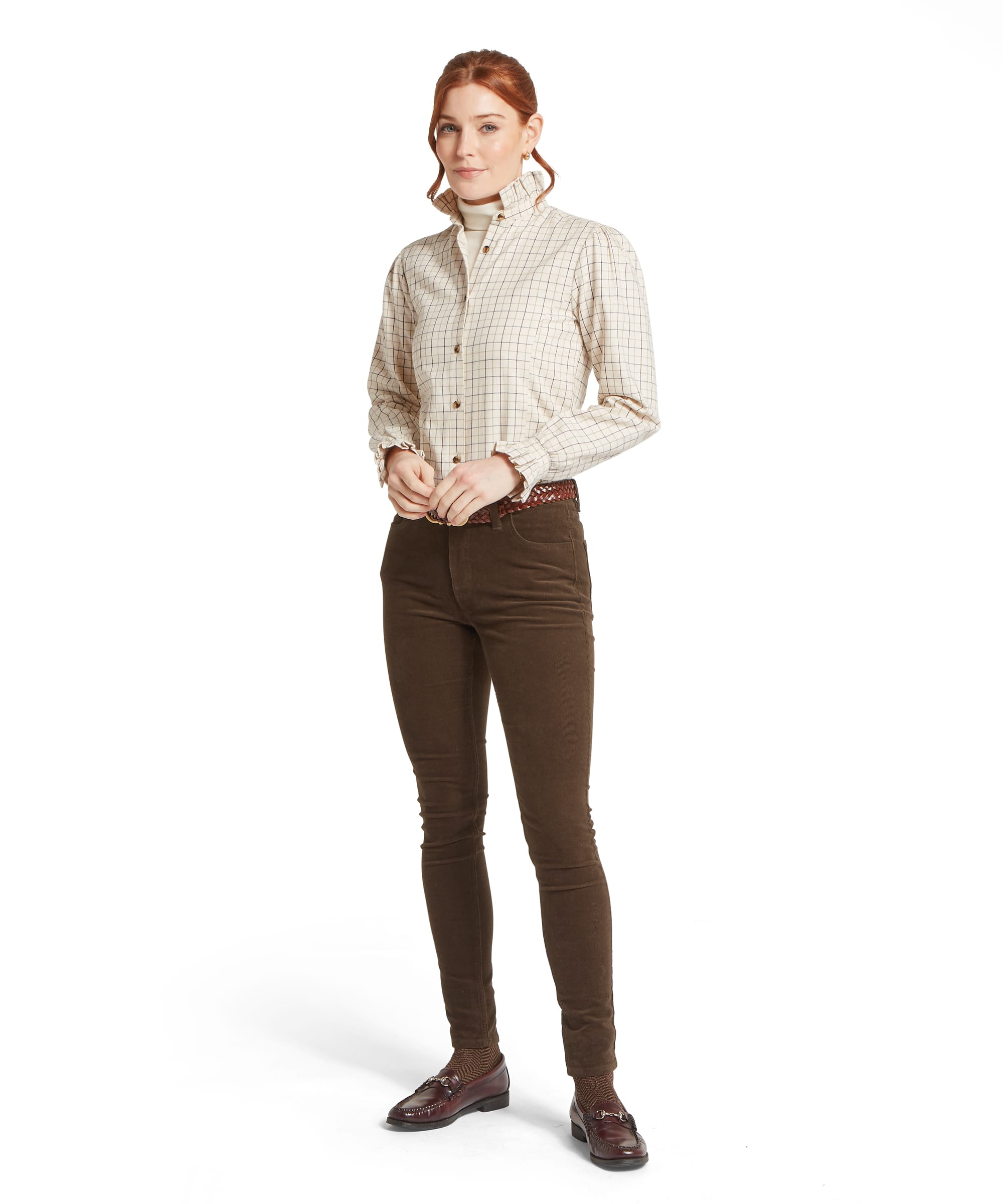Full-length view of the Women's Schöffel Sheringham Shirt in cream tattersall check, worn open over a cream roll neck as a layer, with slim dark olive cord trousers, a woven leather belt and burgundy horsebit loafers.