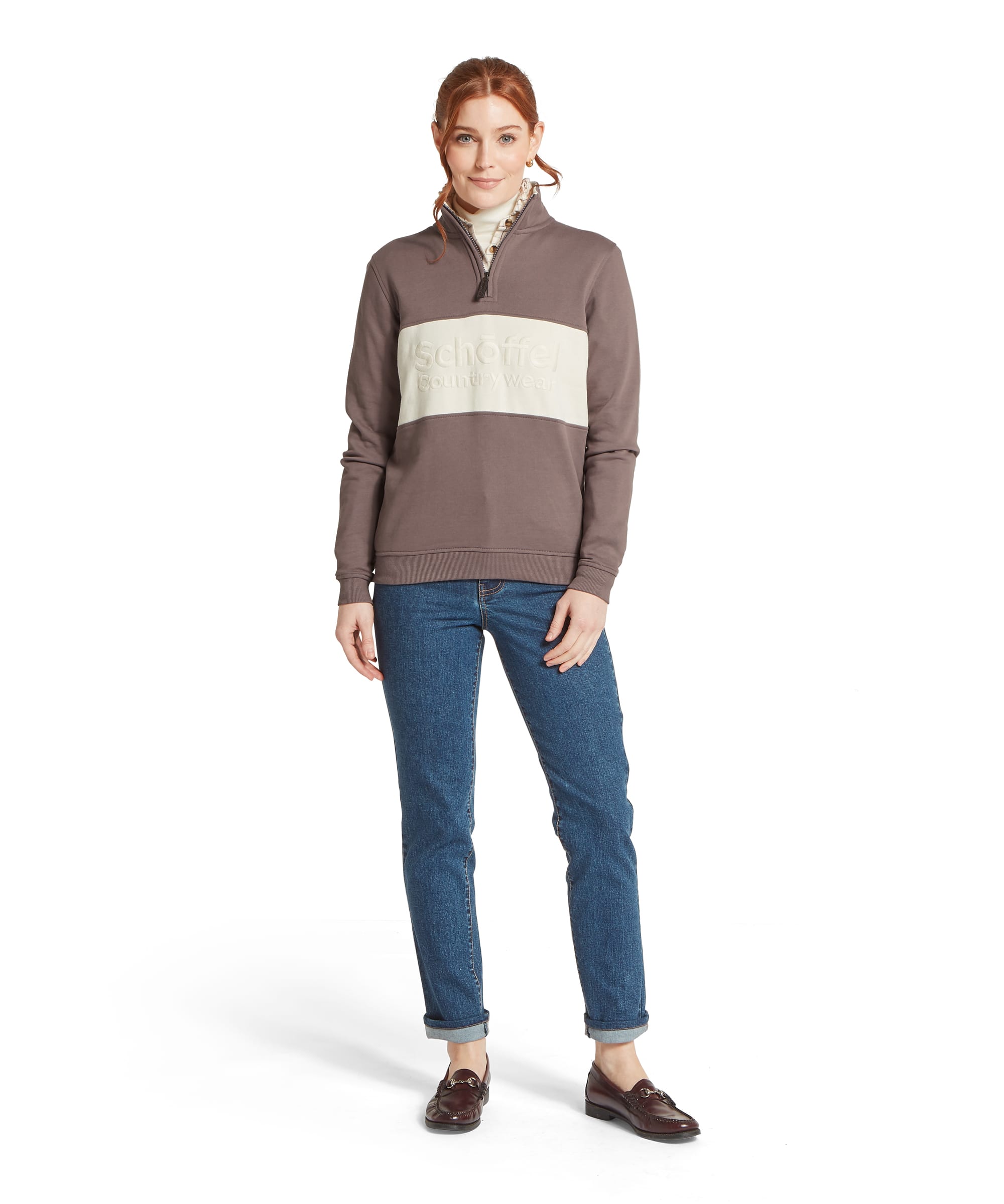 Full-length view of a woman wearing the Schöffel Women's St Agnes Quarter-Zip Sweatshirt in Mink, featuring the tonal embossed Schöffel Country Wear logo on a contrast cream chest panel, layered over a cream roll neck and styled with blue jeans and burgundy horsebit loafers