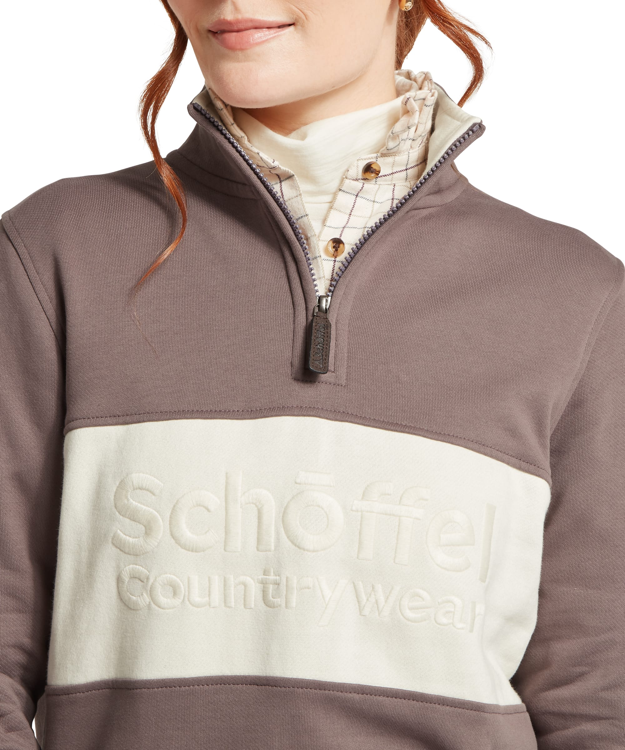Close-up chest and zip detail of the Schöffel Women's St Agnes Sweatshirt in taupe brown, showing the raised cream colourblock panel with embossed Schöffel Countrywear logo, quarter-zip funnel neck with branded Schöffel zip pull, and layered windowpane-check collar beneath