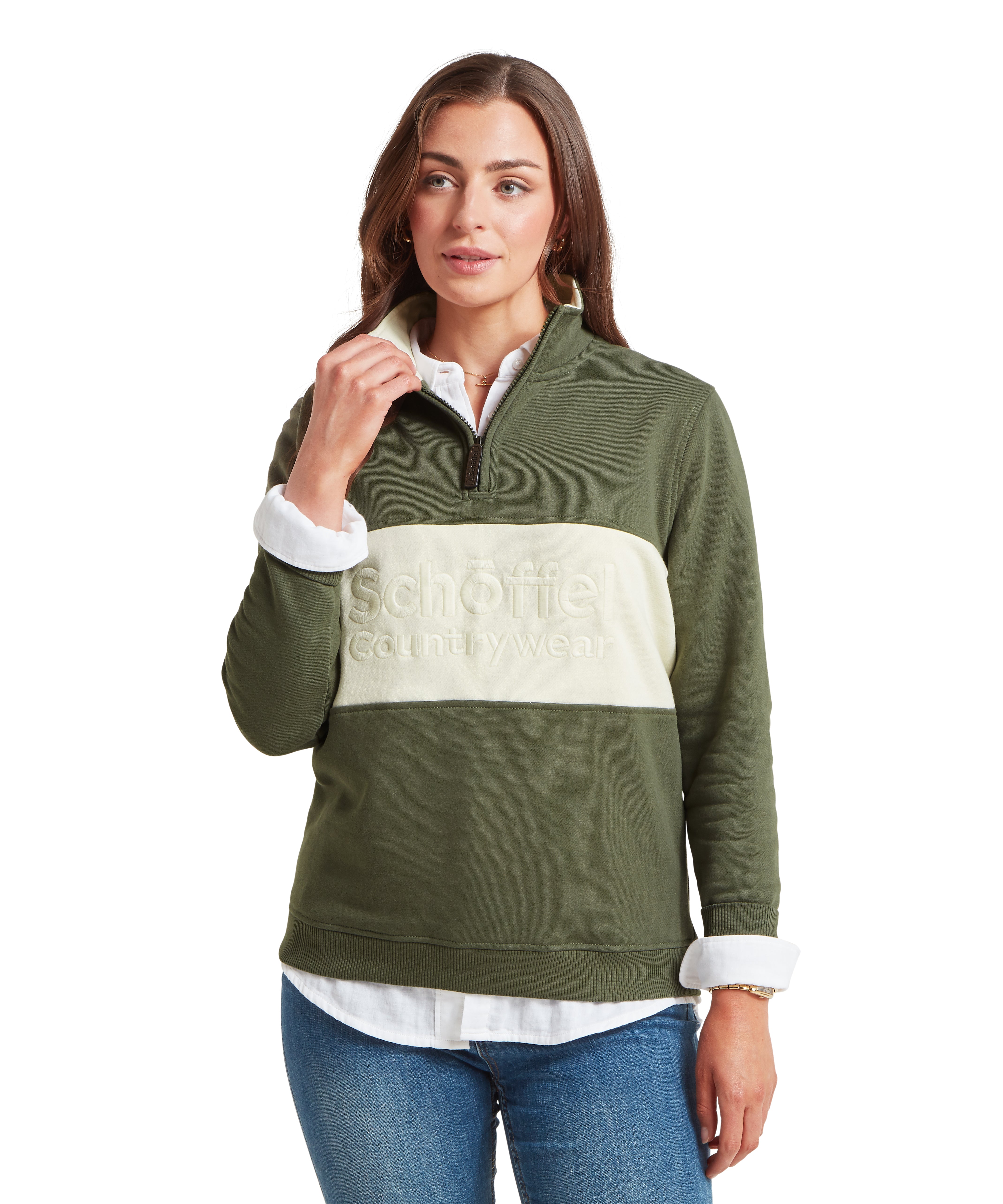 Woman wearing a green and cream quarter zip sweatshirt featuring a Schöffel countrywear logo, paired with blue jeans and a white shirt.  The woman is looking to the side with one arm by her side and the other adjusting her collar.