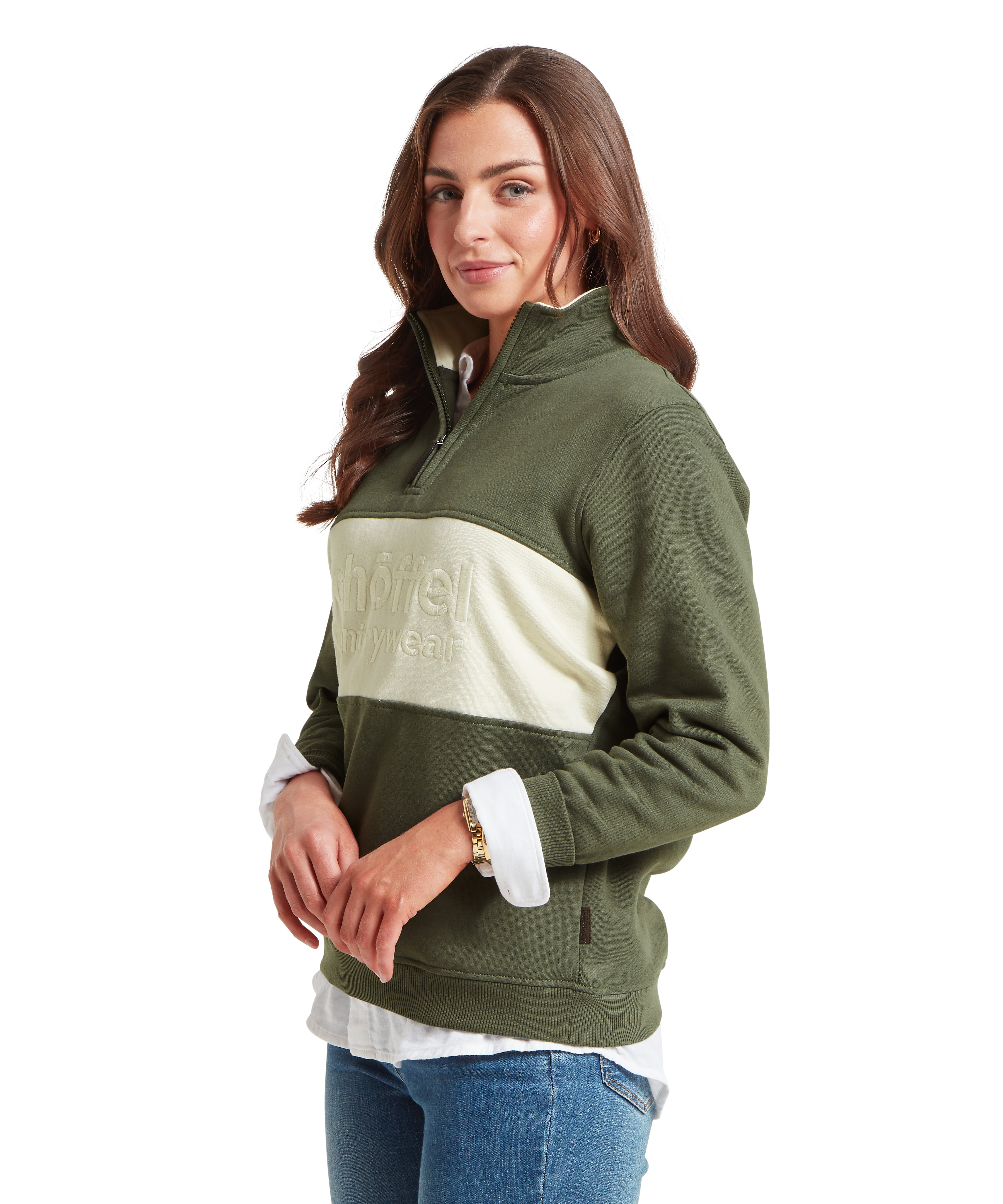 Studio portrait of a woman with long wavy brown hair wearing a dark green quarter zip sweatshirt. The sweatshirt features a cream-colored horizontal chest panel with "schöffel countrywear" embossed on it.