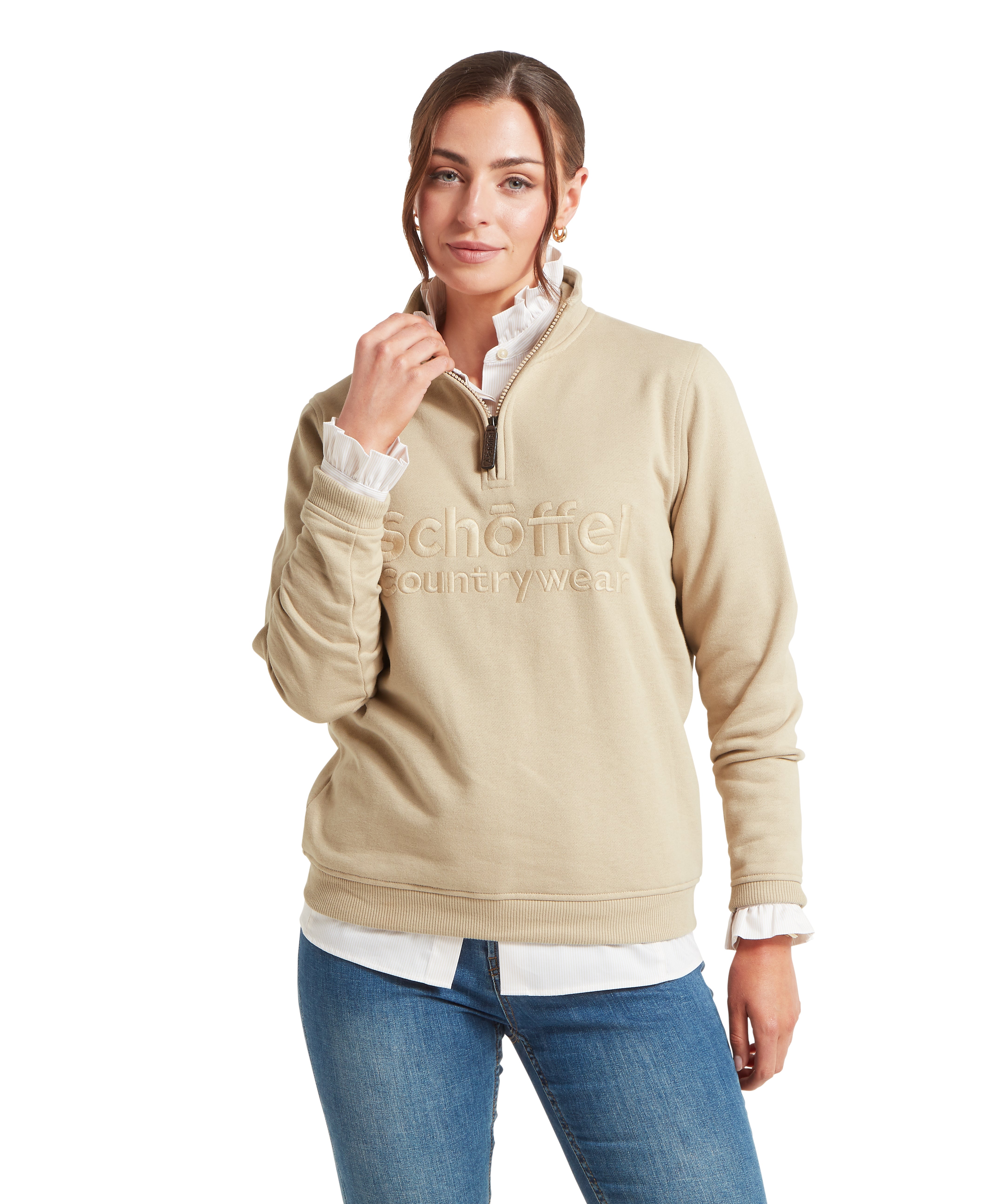 Woman wearing a beige sweatshirt with a Schöffel countrywear logo paired with a white shirt and blue jeans against a white background