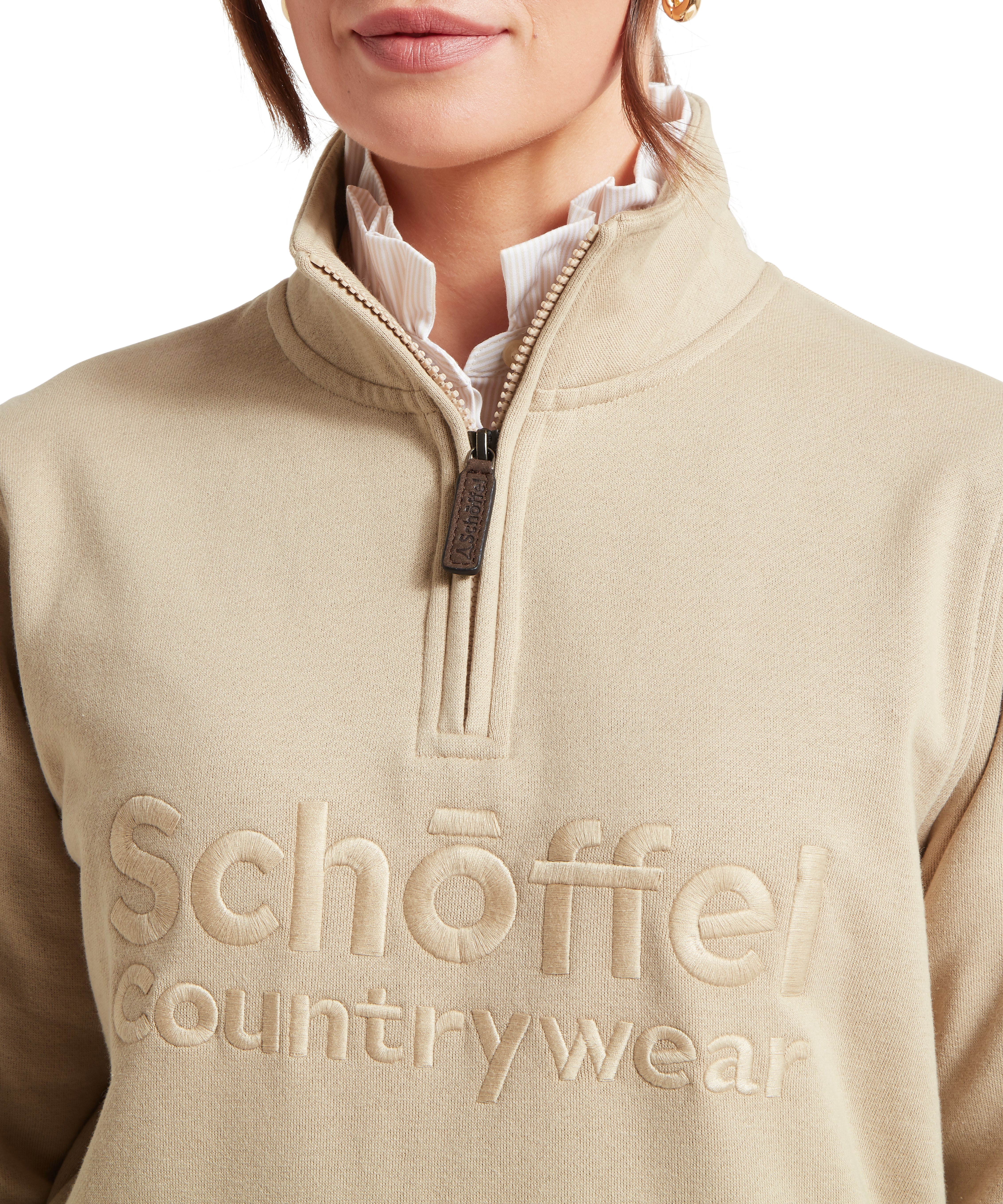 Close up of a woman wearing a beige quarter zip sweatshirt with a Schöffel Countrywear logo on against a white background
