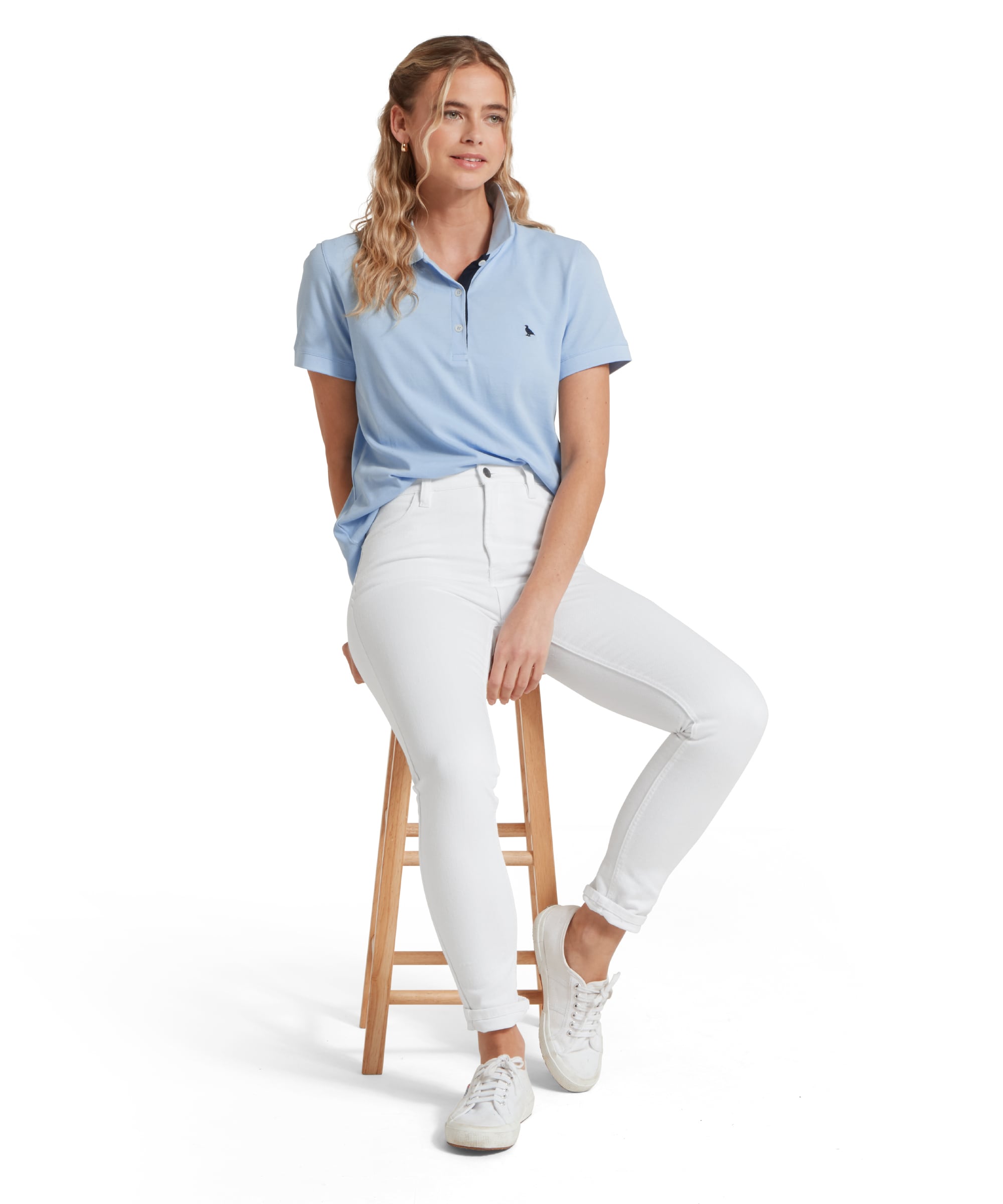 Schöffel Women's St Ives Polo Shirt in pale blue, styled with white jeans and trainers, model seated on wooden stool