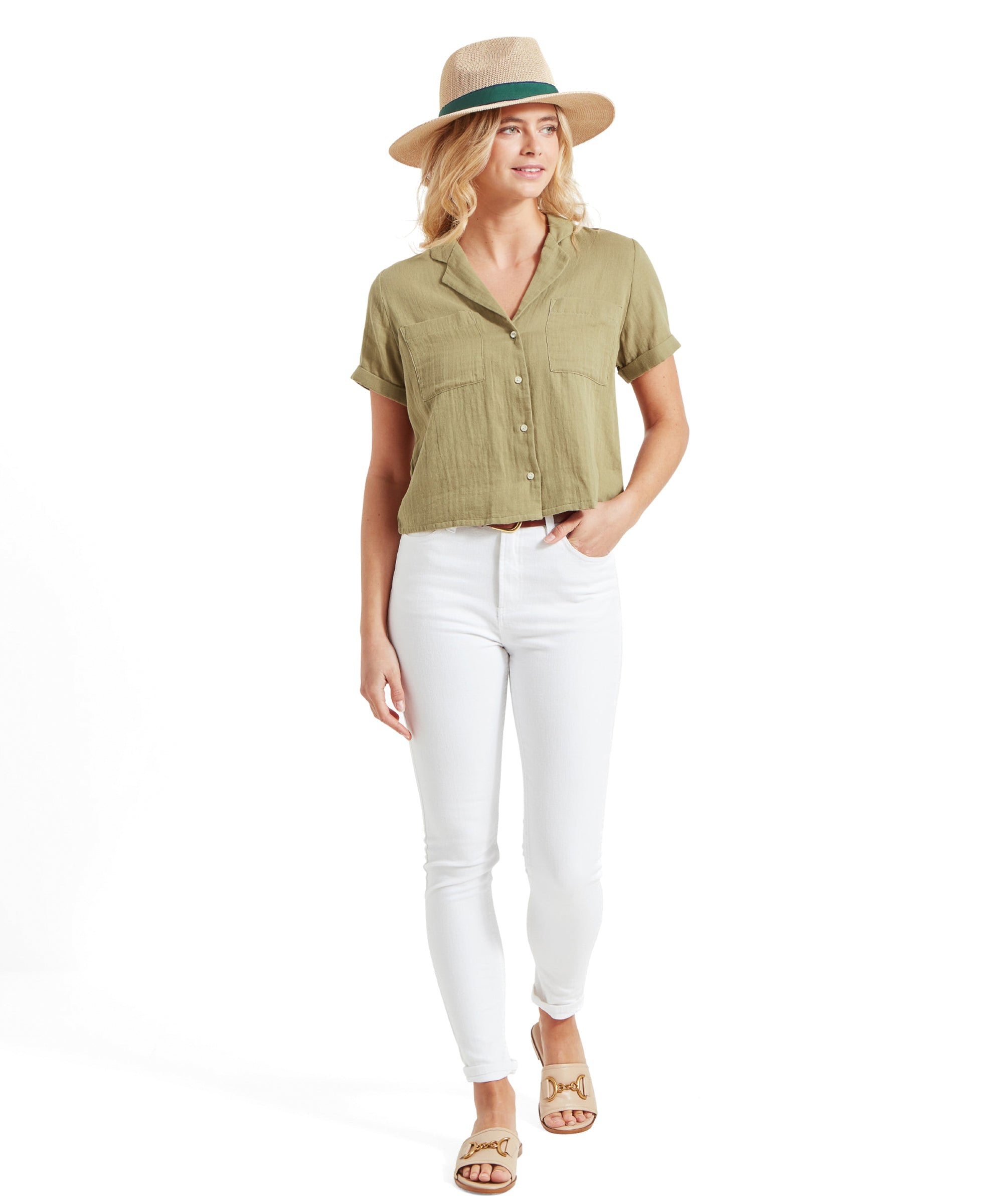 The model adds a hat to her outfit, wearing the Schöffel Summerfield Shirt for Women in Green. She pairs it with white jeans, beige sandals, and a straw hat with a green band. She poses with one hand holding the hat, looking content and stylish.