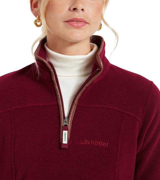 Close-up of a woman wearing a deep burgundy Schöffel quarter-zip fleece pullover, featuring a contrast mauve trim along the zip placket and collar. Layered over a cream turtleneck. The Schöffel logo is embroidered on the chest. The model wears gold hoop earrings.