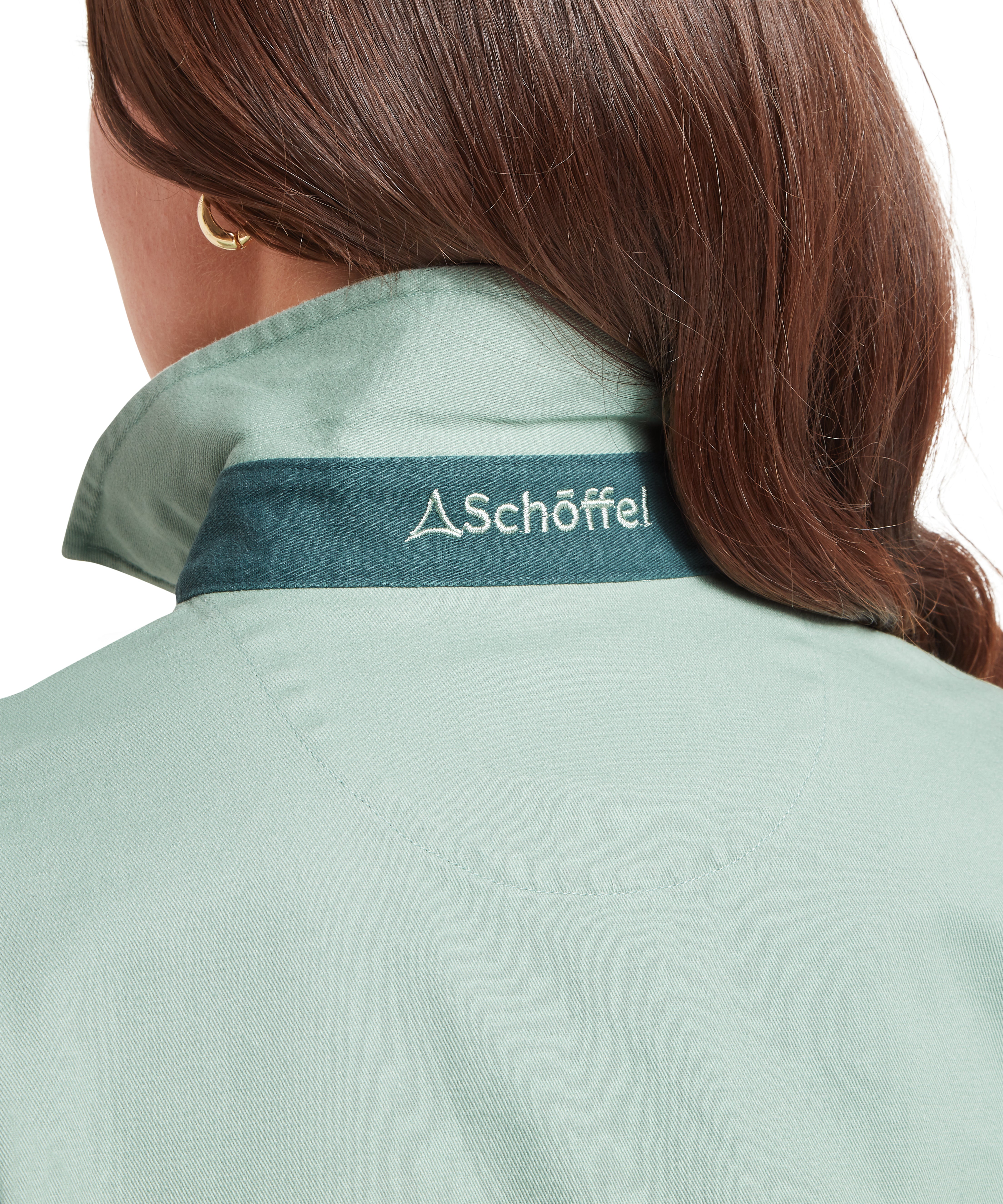 Close up of a woman wearing green deck shirt, showing a dark green accent and Schöffel logo.  The woman is facing backwards with her hair swept to one side.