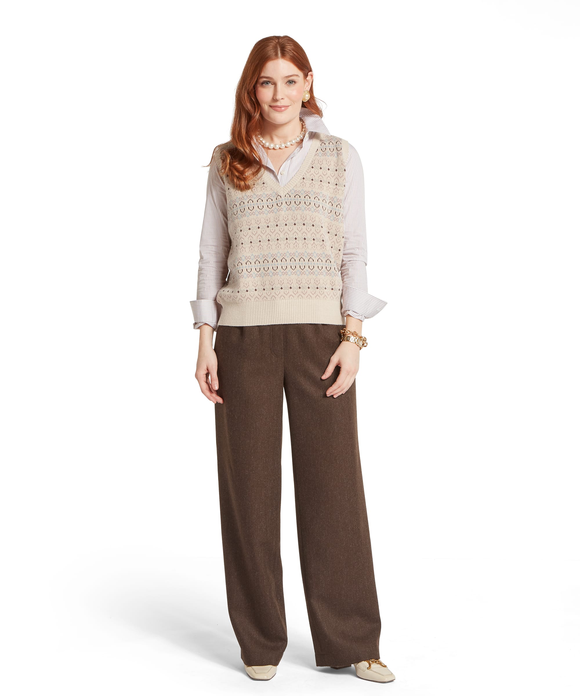 Full-length view of the Women's Schöffel Tresness Fair Isle Vest in oatmeal cream with a V-neck and all-over Fair Isle pattern in dusky pink, blue and brown, layered over a pink stripe shirt with wide-leg brown tweed trousers, a pearl necklace and cream horsebit loafers.